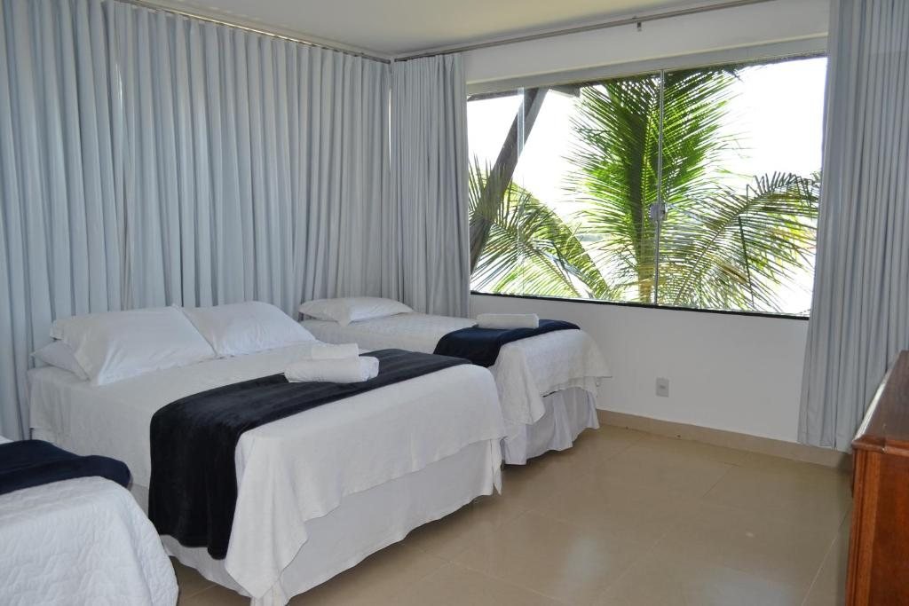Quarto com camas de roupa branca, mantas azul-marinho, cortinas claras e janela ampla